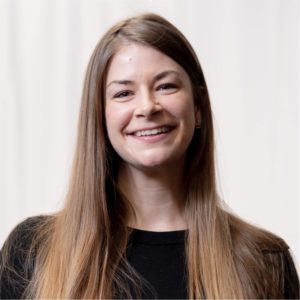 Michelle Dugger is featured in this portrait photograph; she is wearing a round neck black top. Michelle has long brown straight hair and she is smiling at the camera. The background is plain cream color.