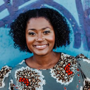 Candy Zollicoffer is featured in this portrait photograph, she is wearing a round neck gray printed blouse with big red, brown, and white. She is smiling at the camera and has ear-length black curly hair.