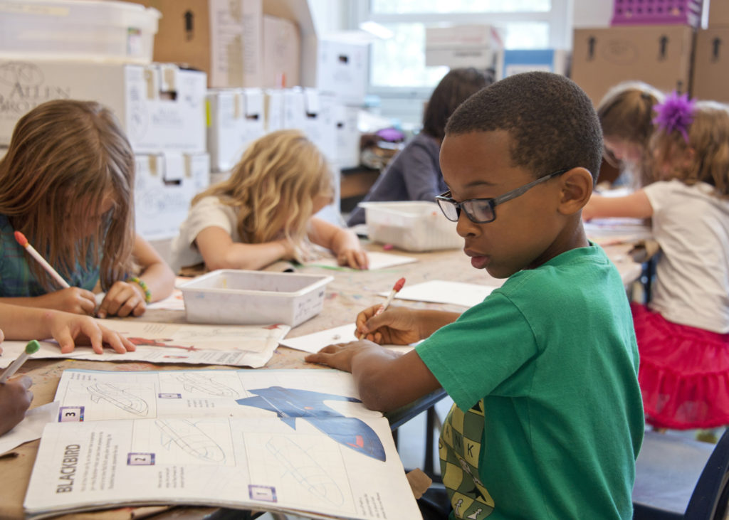The photograph is focused on a Black child in a classroom wearing glasses and a green t-shirt sitting at a table writing on a piece of paper as he looks down on an open magazine on the table, next to him other children also doing the same kind of work. In the background are multiple file boxes surrounding the room.