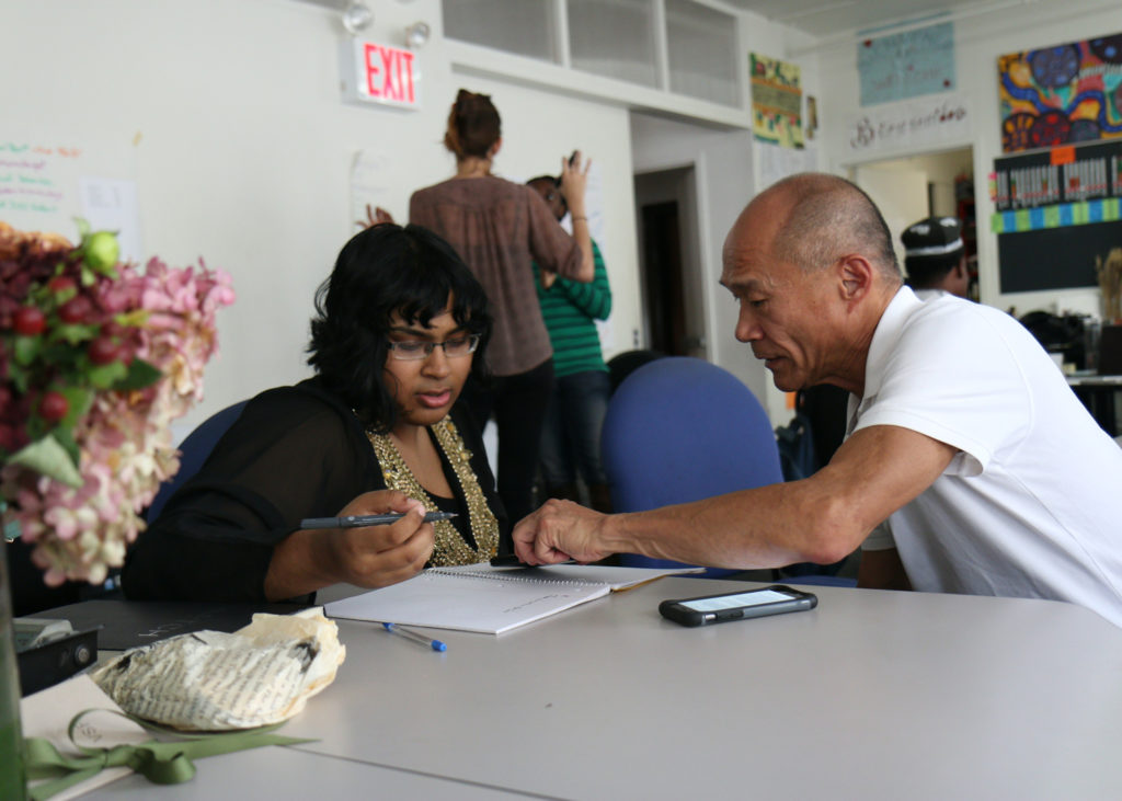 A Black woman seated at a table holding a pen on her right hand listening to an asian elder man seated next to her pointing at something on her notebook. She is wearing a brown top and a black jacket, he is wearing a white polo. The background is a room with white walls, two women are standing and talking, their faces are not visible. There is a flower base on the bottom corner on top of the table.