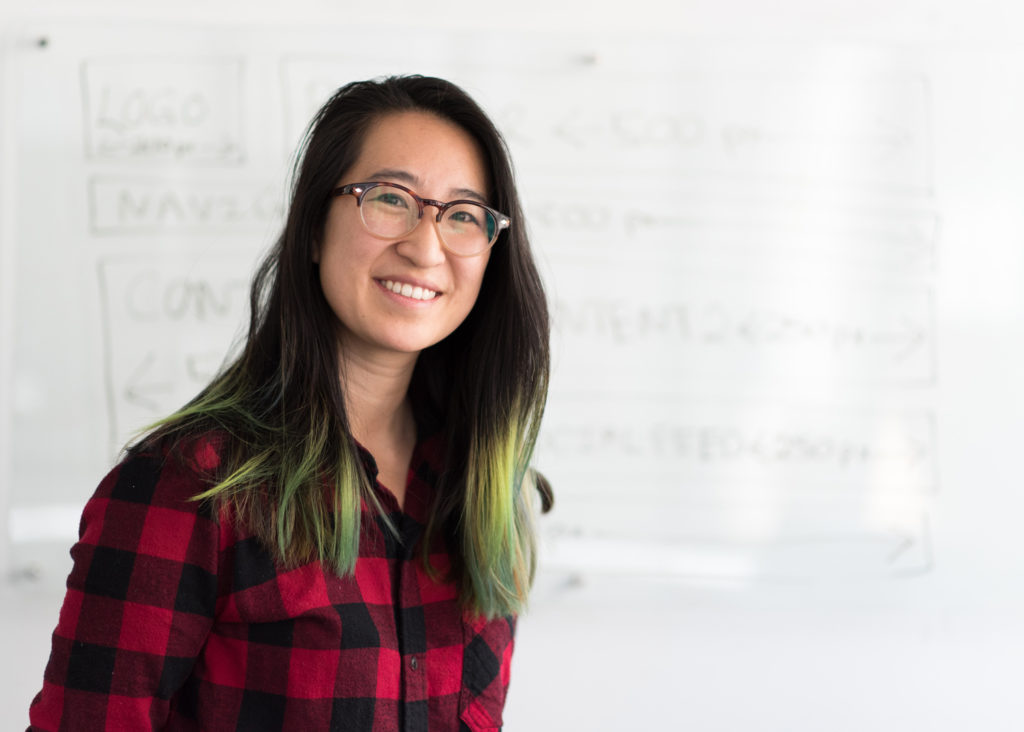 A young Asian woman with long black straight hair on the top and green ends wearing oval glasses and a tartar plaid black and red wool shirt. She is smiling to the camera, in the background a white board with faded words inside rectangles.