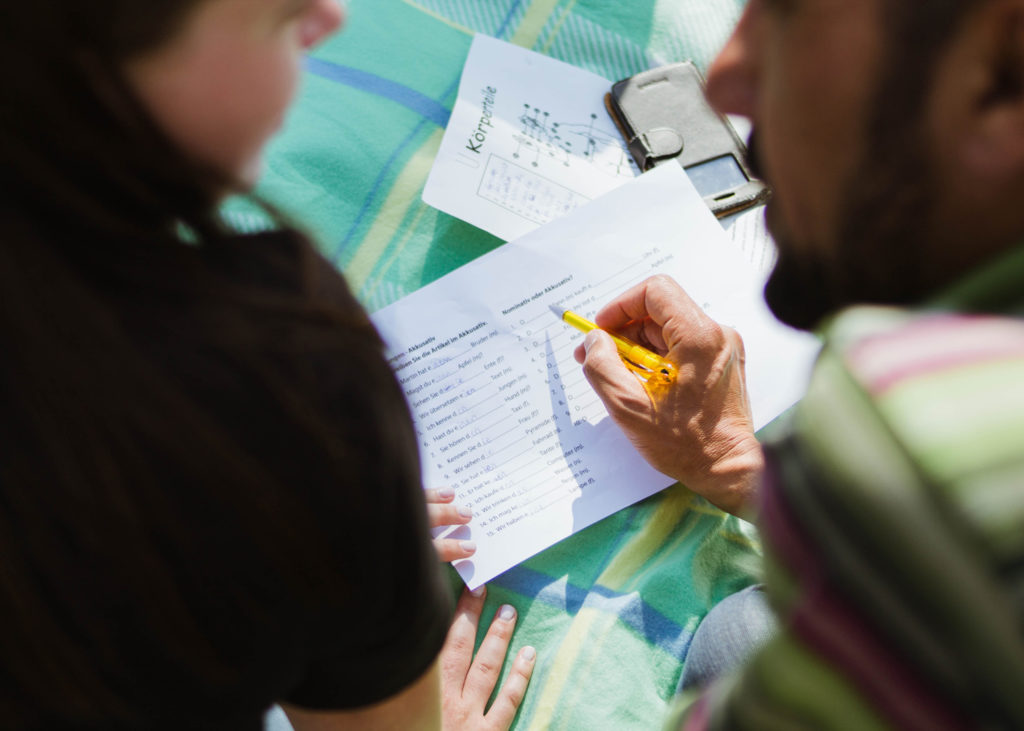 A photograph taken from the top shows a brown man with a long stubble beard, he is wearing a pink and green striped shirt and he is holding a pencil on top of a written paper sheet looking at a white woman with long black hair wearing a black t-shirt. The woman is looking at the man and she is holding her body by placing her hands over a tartar plaid green, yellow and blue blanket on the ground.