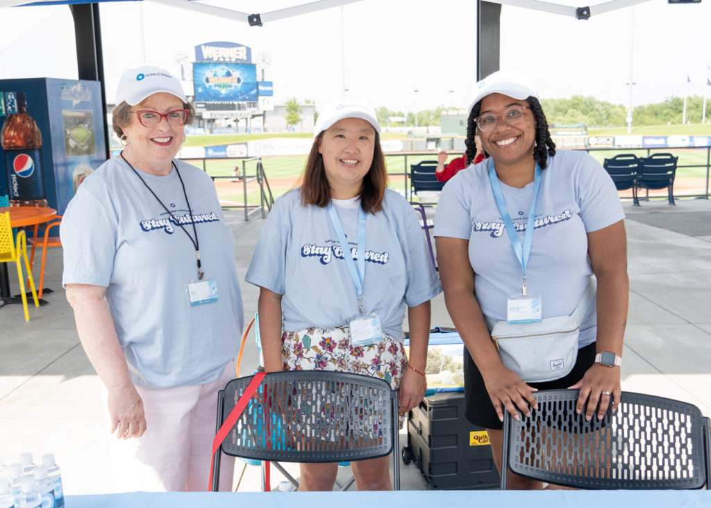Two World Speaks' volunteers and The Executive Director Leah standing next to each other wearing World Speaks’ merchandise (hats and t-shirts) smiling at the camera behind gray chairs at an event. The background is an outdoor event at a local stadium.