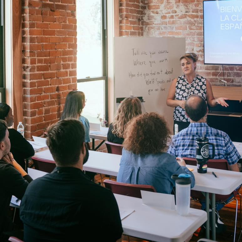 La fotografía muestra una de las lecciones presenciales de uno de los cursos comunitarios. La foto está tomada desde la parte de atrás de la clase, la instructora de World Speaks Ginny, está parada en frente de la clase gesticulando con sus manos a la vez que enseña español a un grupo de estudiantes sentados detrás de 3 filas de mesas blancas rectangulares. Al lado de Ginny, una pizarra blanca con conceptos escritos por la instructora, parte de una pantalla digital se puede ver detrás de Ginny. El salón de clase tiene paredes de ladrillo y dos ventanas grandes rectangulares en el lado izquierdo. 
