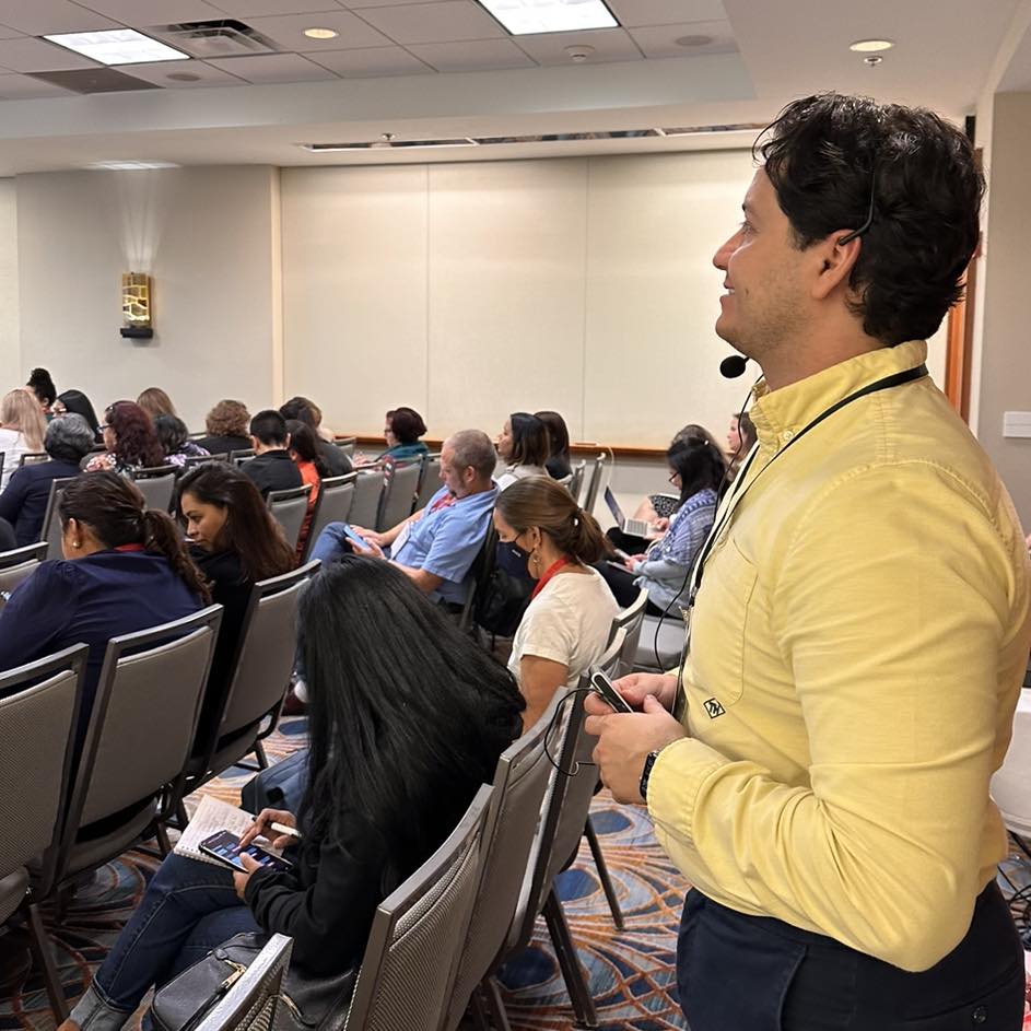 The photograph features World Speaks Language Accessibility Manager Arturo performing interpreting services at a conference. In the front frame Arturo is standing at the back of a room facing sideways to the camera looking towards a stage not visible in the frame. He is wearing a yellow shirt and black pants. A lanyard is visible around his neck as well as a headset over his head, he is holding a transmitter in his hands. In front of him are two rows of chairs with people sitting on them facing the stage not visible on the frame. On his right side is an aisle followed by four rows of chairs with people sitting on them facing the stage not visible on the frame. The wall at the back of the photograph is plain white, you can see a rectangular wall light on towards the front of the wall.