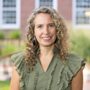 Dr. Melanie Bloom is featured in this portrait photograph. She is wearing a short-sleeved v-neck green top and black pants. Melanie has armpit-length curly blond hair and she is smiling at the camera. The background shows part of a brick-wall building with big white frame windows surrounded by gardens.