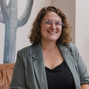 A headshot of Hannah Michelle Bussa, a young white woman with curly brown hair. She is wearing a light green jacket and black shirt underneath. She is smiling through silver glasses. The background has a white wall with a grey cactus painted onto it.