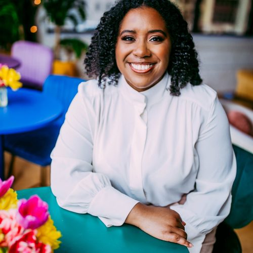 Leah Whitney Chavez, a Black woman, is featured in this photograph. She is wearing a white shirt with long sleeves and is sitting at a teal-colored table. The foreground has pink and yellow flowers, and the background has colorful chairs, tables, and plants. Leah is smiling at the camera, and she has shoulder length curly black hair.