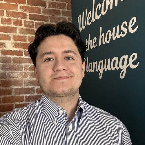 Arturo Aceves Gonzalez is featured in this portrait photograph. He is wearing a blue & white striped collar shirt. Arturo has short wavy dark brown hair, and he is showing a closed-mouth smile. The background shows a bricked wall on the left and a dark painted wall with a round white letter phrase that reads “Welcome to the house of language” on the right.