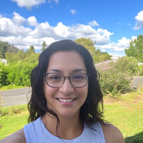 Paulina Campos Guzman, a Latina woman, is featured in this portrait photograph. She is wearing a light gray sleeveless T-shirt, and black oversized oval shaped glasses. Paulina has shoulder length wavy dark brown hair and she is smiling at the camera. The outdoor background shows blue skies with cumulus clouds, a horizontal paved street with different shaped trees on the top side, and a grass area on the bottom side.