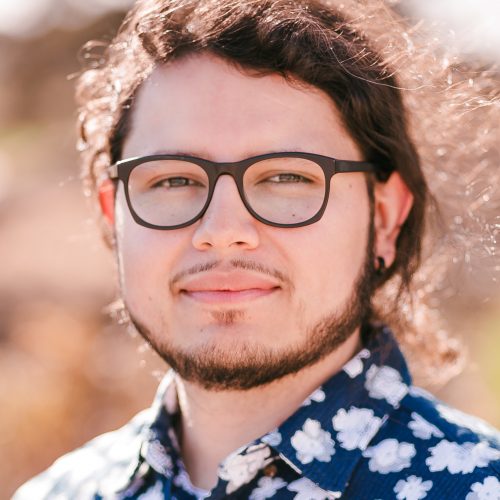 Randy Whitney Chavez is featured in this portrait photograph. He is wearing a blue collar shirt printed with white flowers and black square shaped glasses. Randy is showing a closed-mouth smile, he has a short Brett beard, a pencil mustache and long curly brown hair on a low ponytail style. The outdoor background is blurry in light and dark brown tones.