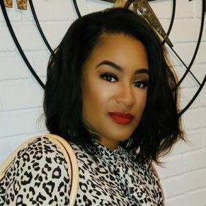 Jasmine Elmore is featured in this portrait photograph, and prefers the pronouns “she, her, hers”. She is an African-American female with brown eyes and shoulder length black hair. Her body is standing sideways, and her face is turned to the direction of the camera. She is wearing a light and dark brown animal print shirt and a handbag which only shows the purse straps visible over her right shoulder. The background shows a white color brick wall and the bottom of a round metal wall decoration.